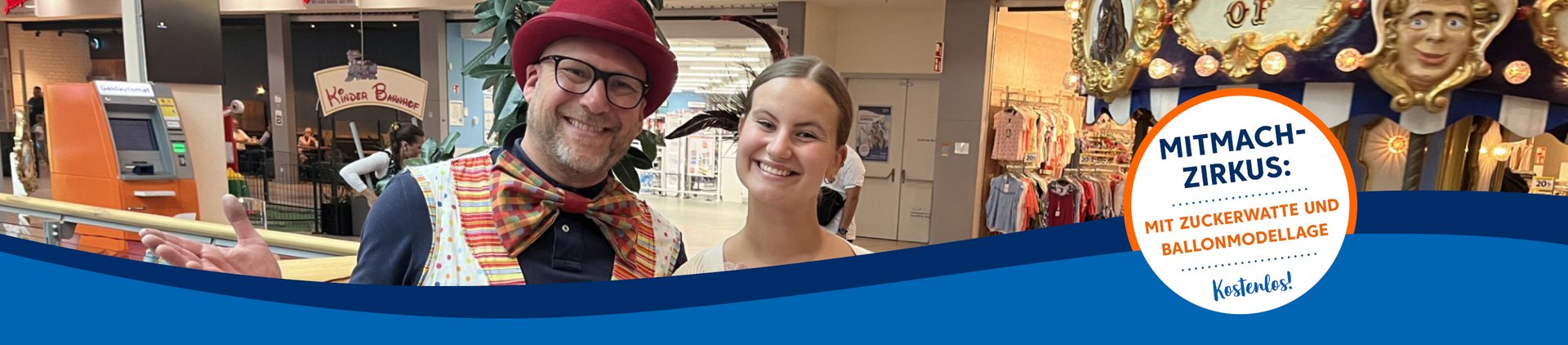 Mann im bunten Outfit und Frau stehen lächelnd im Einkaufszentrum, Werbeplakat für Mitmach-Zirkus im Hintergrund.